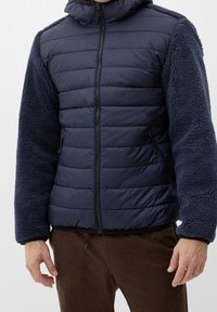 Uomo in giacca trapuntata blu navy con maniche in pile testurizzato e pantaloni marroni, in piedi di fronte a uno sfondo semplice.