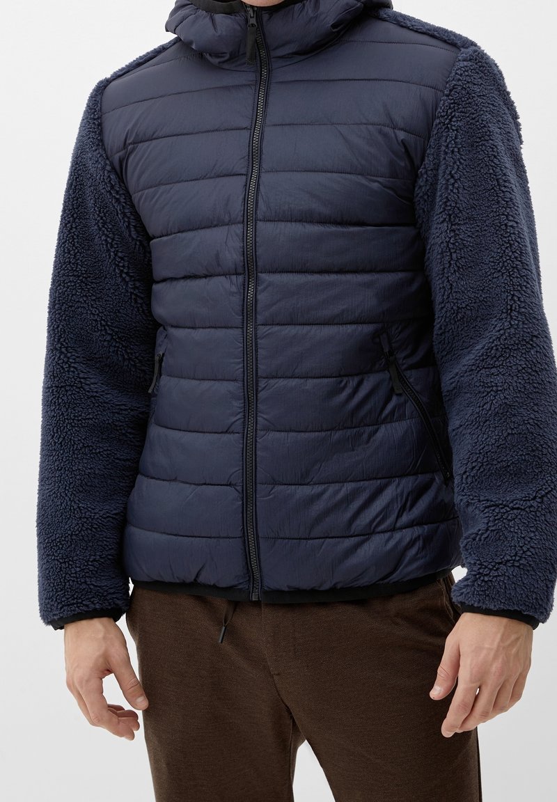 Uomo in giacca trapuntata blu navy con maniche in pile testurizzato e pantaloni marroni, in piedi di fronte a uno sfondo semplice.
