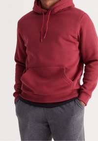 Burgundy huvtröja med dragsko i huvan, kängurufickor och ribbade manschetter. Bärs över mörkgråa sweatpants. Slät textur, avslappnad design.