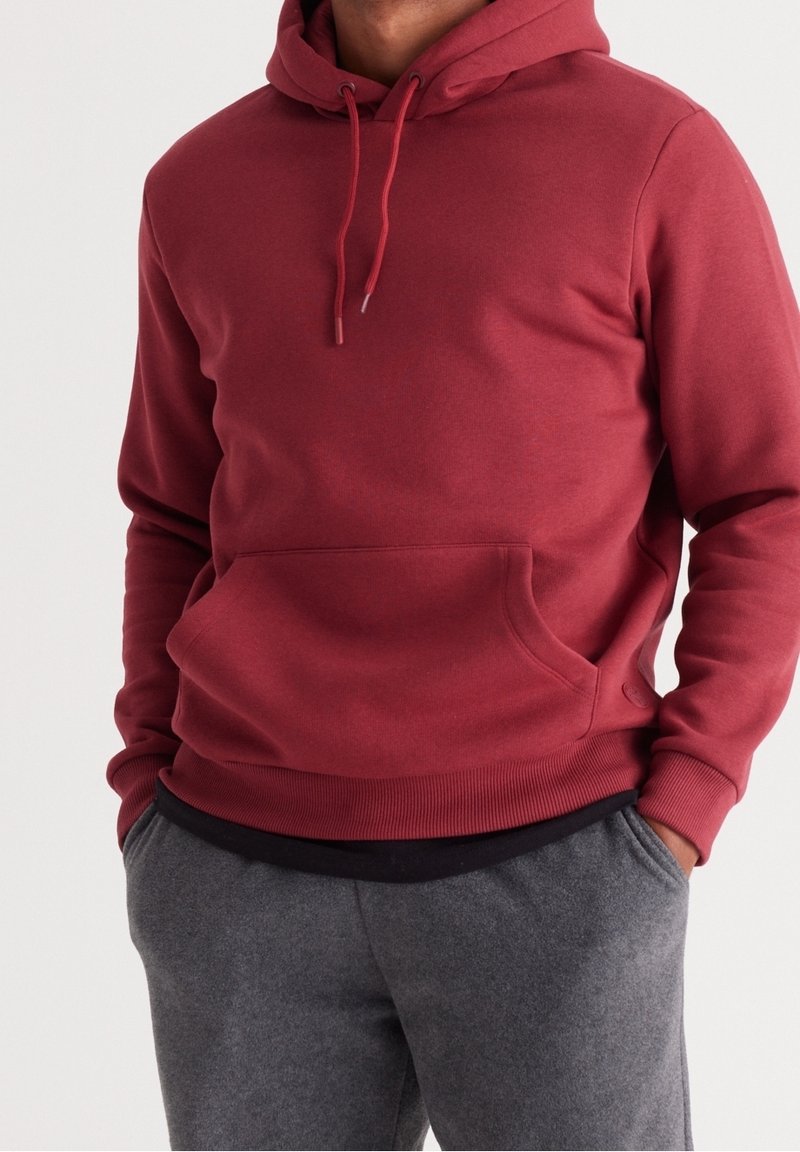 Burgundy huvtröja med dragsko i huvan, kängurufickor och ribbade manschetter. Bärs över mörkgråa sweatpants. Slät textur, avslappnad design.