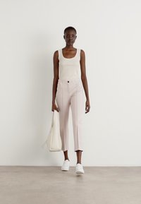 Beige tanktop, lichtroze op maat gemaakte broek met voornaaddetails en witte sneakers. Het model draagt een lichtbeige tas. Minimalistisch ontwerp.