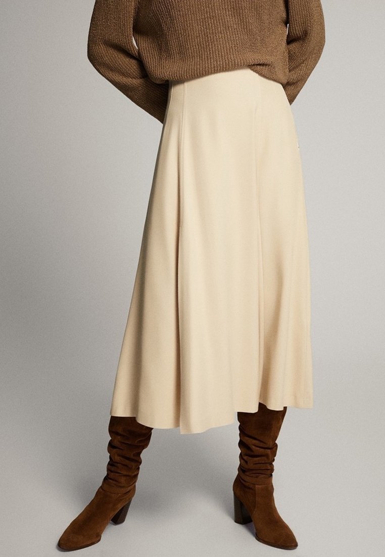 Personne portant une jupe midi beige en ligne A, des bottines en daim brun décontractées à talons, et un pull en maille épaisse marron avec les mains dans le dos.