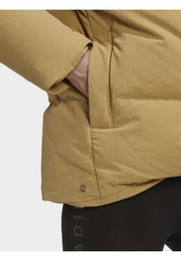Main dans la poche d'une veste matelassée marron portée sur un legging noir avec un texte discret sur la jambe, visible de la taille jusqu'au milieu de la cuisse sur un fond neutre.