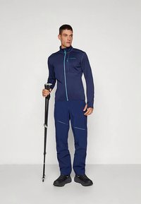 Tumesinine välisjope ja sobivad püksid, millel on tõmblukuga sulgemine, hele sinised aktsendid ja sile, tehniline kangas. Kasutaja hoiab käepidemega matkakeppi.