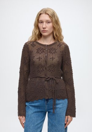 Jeune femme aux longs cheveux blonds porte un cardigan en maille marron boutonné avec des motifs floraux et un jean bleu sur un fond uni.