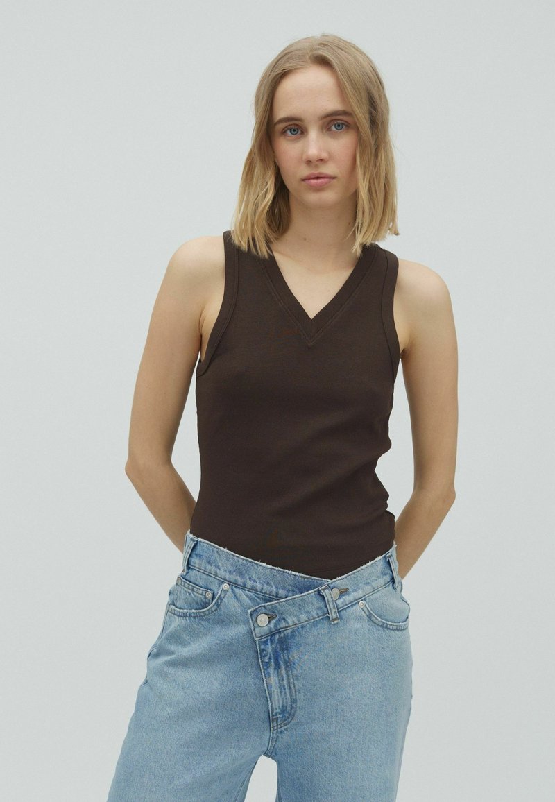 Jeune femme aux cheveux blonds portant un haut sans manches marron foncé et un jean bleu oversize sur un fond gris clair uni.