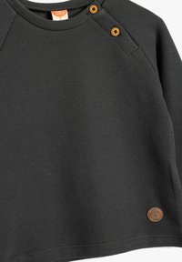 Camisa de bebé de manga larga de color gris oscuro con un diseño acanalado, dos botones naranjas en el hombro y un pequeño parche de cuero redondo cerca del dobladillo.