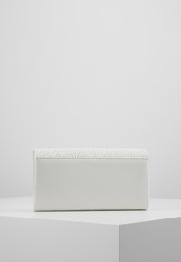 Pochette rectangulaire blanche avec un corps en tissu lisse et des détails en dentelle texturée le long du bord supérieur. Fermeture éclair visible à l'arrière.