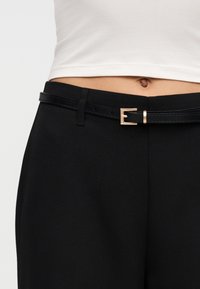 Κοντινό πλάνο της μέσης που φοράει μαύρο παντελόνι με λεπτή μαύρη ζώνη και λευκό κοντό τοπ, με εκτεθειμένο δέρμα και ομφαλό.