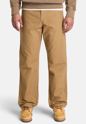 Mann trägt beige gerade geschnittene Hosen, braune Schnürstiefel und einen beigen Pullover mit weißem Unterhemd vor weißem Hintergrund.