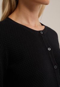 Cardigan nero texturizzato con scollo rotondo, bottoni frontali e motivo a nido d'ape. Tessuto morbido e forma aderente.