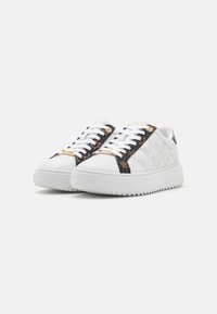 Sneaker bianca con texture trapuntata, accenti neri sui lati, lacci piatti, suola spessa bianca e dettaglio del logo vicino alla linguetta.