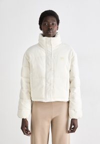 Veste matelassée coupée en tissu côtelé crème avec col montant, fermeture éclair à l'avant et poches latérales. Texture lisse avec un branding minimaliste.