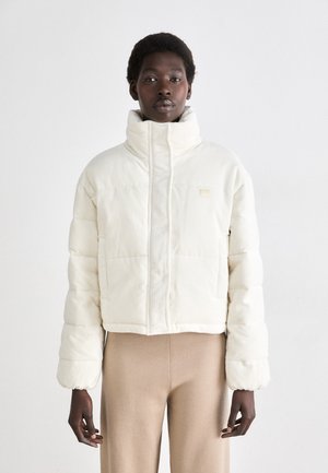 Veste matelassée coupée en tissu côtelé crème avec col montant, fermeture éclair à l'avant et poches latérales. Texture lisse avec un branding minimaliste.