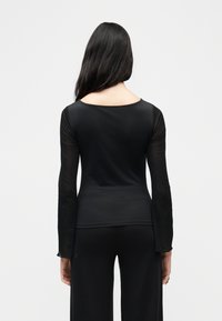 Γυναίκα με μακριά σκουρόχρωμα μαλλιά φορεί στενή μαύρη μπλούζα με μακριά μανίκια και χαλαρό μαύρο παντελόνι, στέκεται απέναντι σε ένα απλό ανοιχτό γκρι φόντο.