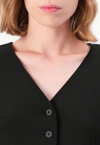 Femme à la peau claire et aux cheveux blonds portant un haut noir à col en V boutonné, dévoilant le décolleté et les clavicules sur un fond uni.