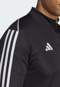 Chaqueta deportiva negra con cuello alto, que presenta tres rayas blancas en el hombro y un logo blanco de Adidas en el pecho.