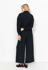 Combinaison noire à manches longues et jambes larges, avec une taille ceinturée et une texture lisse, associée à des chaussures noires brillantes.