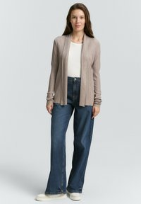 Strickjacke in hellbeige mit vertikaler Rippung, über einem weißen Oberteil getragen. Weite blaue Jeans und cremefarbene Sneaker runden das Outfit ab.