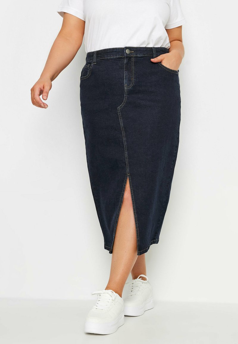 Falda midi de mezclilla oscura con una abertura frontal, cintura alta y dos bolsillos. Presenta costuras amarillas y se combina con zapatillas blancas.