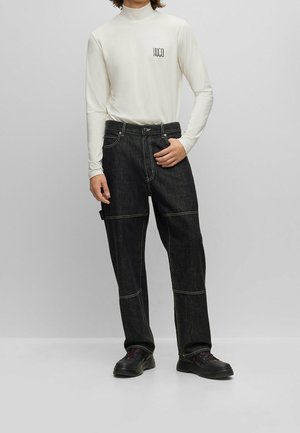 Bílá tričko s dlouhým rukávem a stojáčkem, s malým černým logem na hrudi, doplněné tmavými džínami s kontrastním stehem a černými botami.