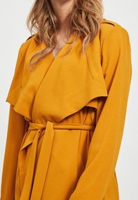 Płaszcz trencz w kolorze musztardowej żółci, z drapowanym kołnierzem, taliowany paskiem i gładką fakturą materiału. Szczegółowe przeszycia wzdłuż krawędzi.