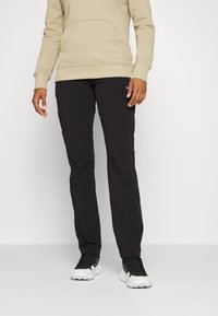 Svarta, avsmalnande byxor tillverkade av lätt material, med en slät yta och diskret branding, kombinerade med texturerade svarta träningsskor.