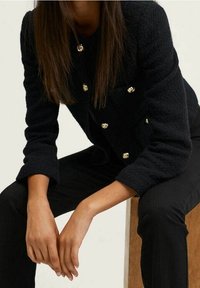 Femme portant une veste noire texturée avec des boutons dorés, assise sur un bloc de bois, les mains reposant entre ses genoux.