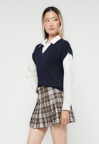 Pull sans manches en tricot bleu marine avec un col en V, porté sur une chemise blanche à col. Associé à une jupe plissée à taille haute en tartan beige et marron.