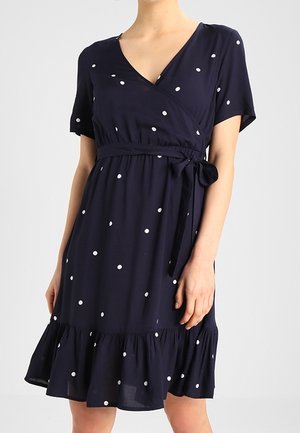 Femme portant une robe bleu marine mi-longue à pois blancs, manches courtes, col en V, ceinture à la taille et ourlet volanté.