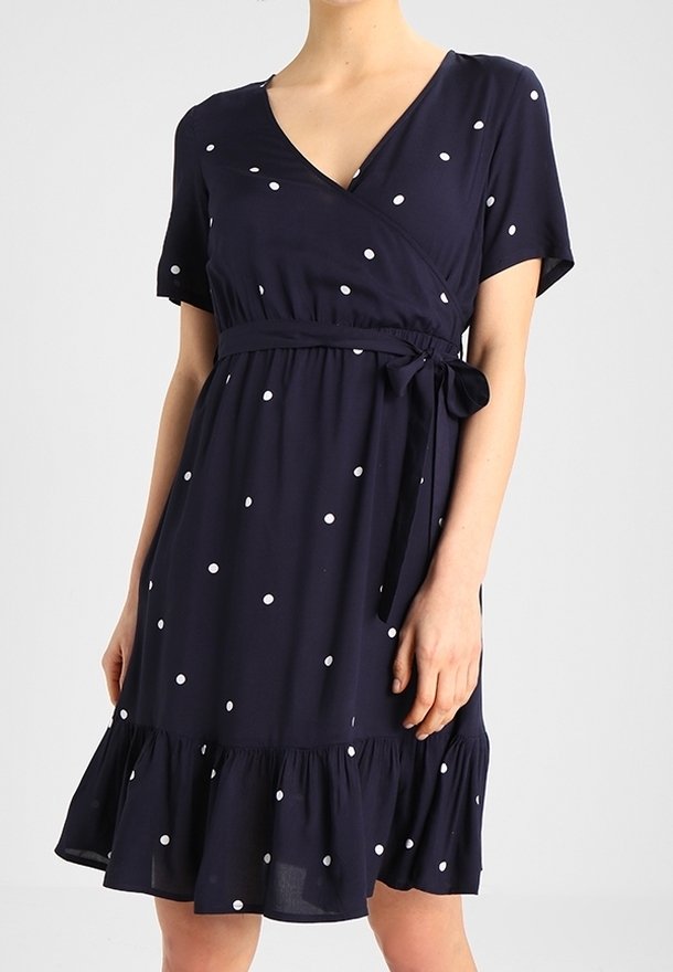 Femme portant une robe bleu marine mi-longue à pois blancs, manches courtes, col en V, ceinture à la taille et ourlet volanté.