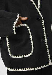 Svart jacka med texturerad tyg, med vit scallopkant längs kanterna och fickorna, samt svarta knappar framtill.