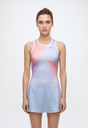 Une femme se tient de face, vêtue d’une robe de tennis sans manches aux nuances dégradées de rose et de lavande, sur un fond blanc uni.
