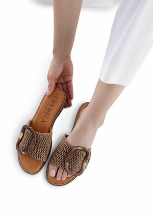 Sandalias marrones tejidas estilo slip-on con una hebilla decorativa grande y plantilla color canela, que cuentan con una suela plana, textura suave y costuras decorativas.