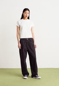Hvid cropped t-shirt med rund halsudskæring, parret med sorte stribede vidde bukser og sorte sneakers. Enkel baggrund med grøn gulv.