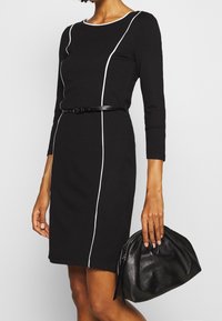Robe noire avec des piping verticaux blancs, manches longues, décolleté rond et une silhouette ajustée, accompagnée d'un sac à main en cuir noir.