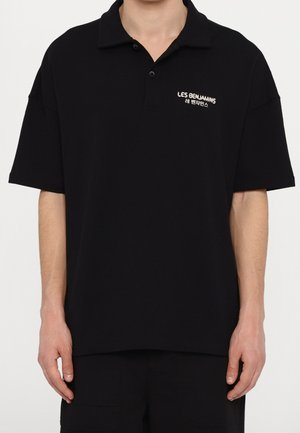 Persona con una camisa polo negra de manga corta con texto blanco "LES BENJAMINS" y caracteres coreanos en el pecho izquierdo.