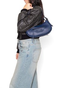 Borsa a tracolla in pelle blu navy con forma curva, superficie texture, chiusura con zip e dettaglio in nappina, indossata sopra una giacca scura e jeans chiari.