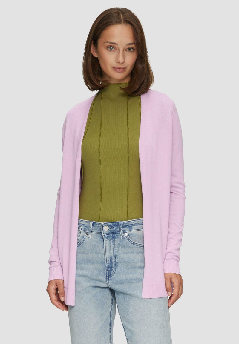 Lichtroze cardigan met een open voorkant en lange mouwen, gecombineerd met een aansluitende groene coltrui. Lichtblauwe high-waisted jeans zijn zichtbaar.