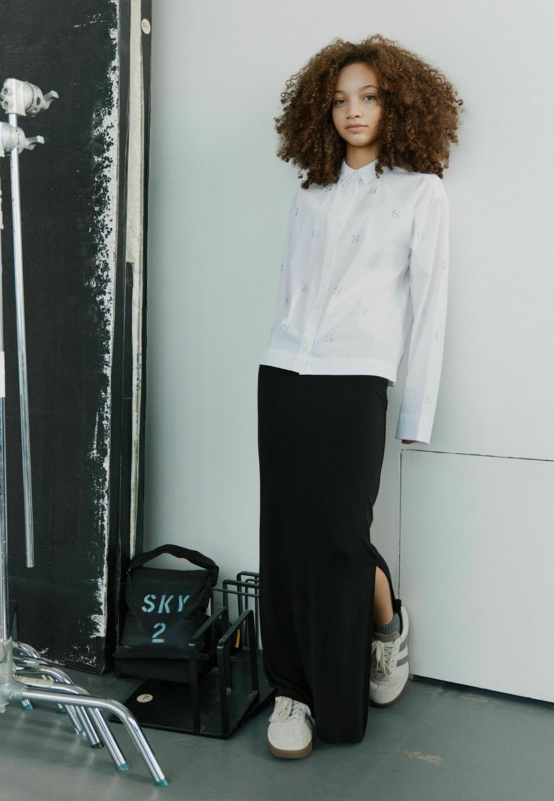 Hvid skjorte med diskret mønster, parret med en lang sort nederdel med en lændeslids. Grå sneakers fuldender looket.