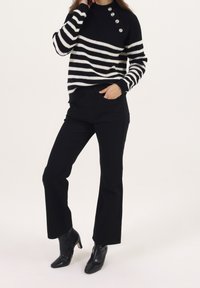 Femme portant un pull rayé noir et blanc avec des boutons argentés, un pantalon noir et des bottines à talons noires, sur fond blanc.