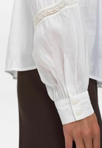 Camicia bianca trasparente a maniche lunghe con dettagli in pizzo e polsini abbottonati, indossata con pantaloni marrone scuro, con focus su braccio e mano.