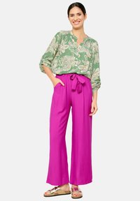 Blusa verde con motivo paisley, con bottone frontale, abbinata a pantaloni magenta a vita alta e gamba larga con dettaglio in vita legato.