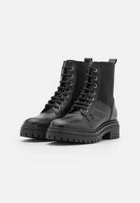 Botas de cuero negras hasta el tobillo con una suela de goma gruesa, que presentan una combinación de superficies lisas y texturizadas, puntera redonda y cordones en la parte delantera.