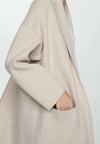 Beige neulotttu neuletakki, jossa avoin edusta, leveät hihat ja suuri tasku vasemmalla puolella. Pehmeä pinta, rento istuvuus ja yksinkertainen muotoilu.