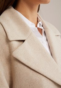 Beige ullblandad kappa med bred krage, jämn textur och strukturerad form, lagd över en vit skjorta med knappar.