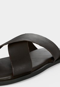 Σανδάλι slide από σκούρο καφέ δέρμα με δύο ευρείες επικαλυπτόμενες λουριά και επίπεδη μαύρη σόλα σε λευκό φόντο.