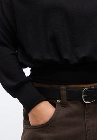 Άτομο που φοράει μαύρο πουκάμισο με μακριά μανίκια τυλιγμένο μέσα σε καφέ παντελόνι με μαύρη ζώ νη, με το χέρι μερικώς στην εμπρόσθια τσέπη.