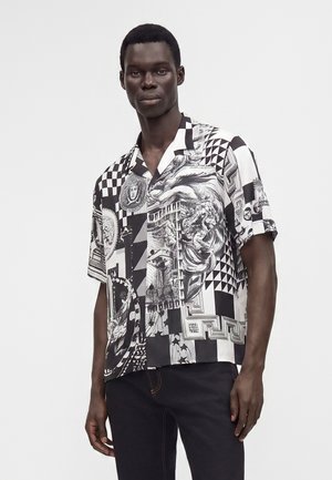 Homme portant une chemise à manches courtes noire et blanche avec des motifs complexes comprenant des visages, des formes géométriques et des motifs d'art classique.