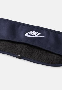 Navy modra flisasta traka za glavo s mehko notranjostjo, ki na sprednji strani nosi izstopajoč beli logotip Nike.